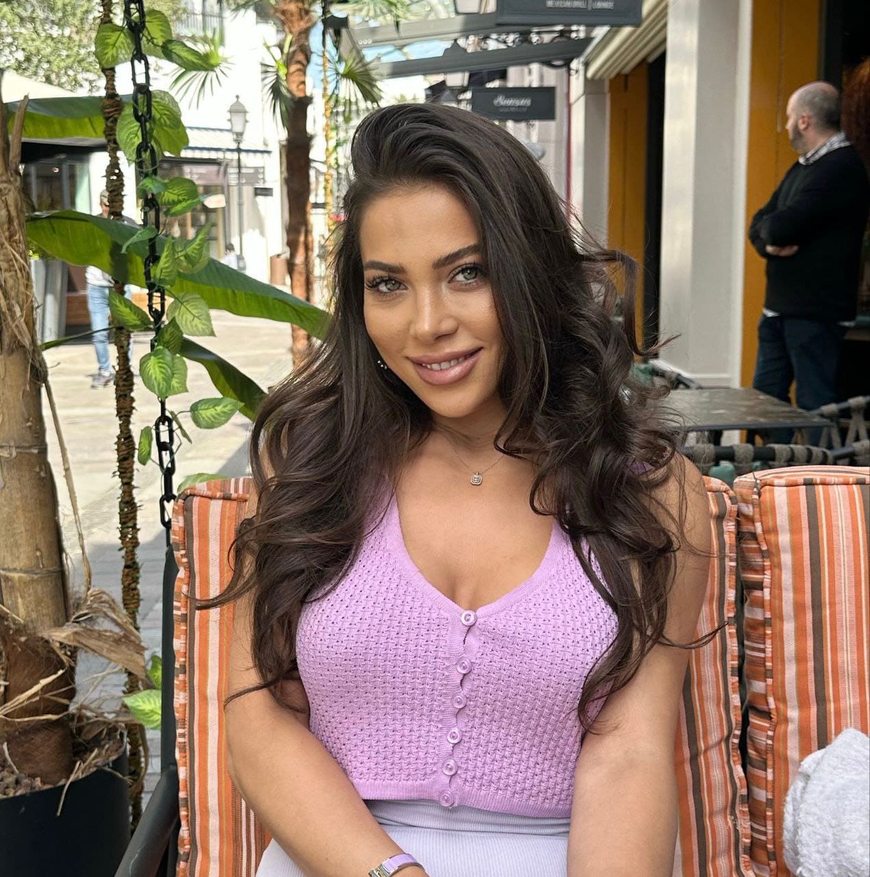Yulya sits in a striped outdoor chair wearing a light purple sleeveless knit top and necklace, with palm trees and a street scene behind her.