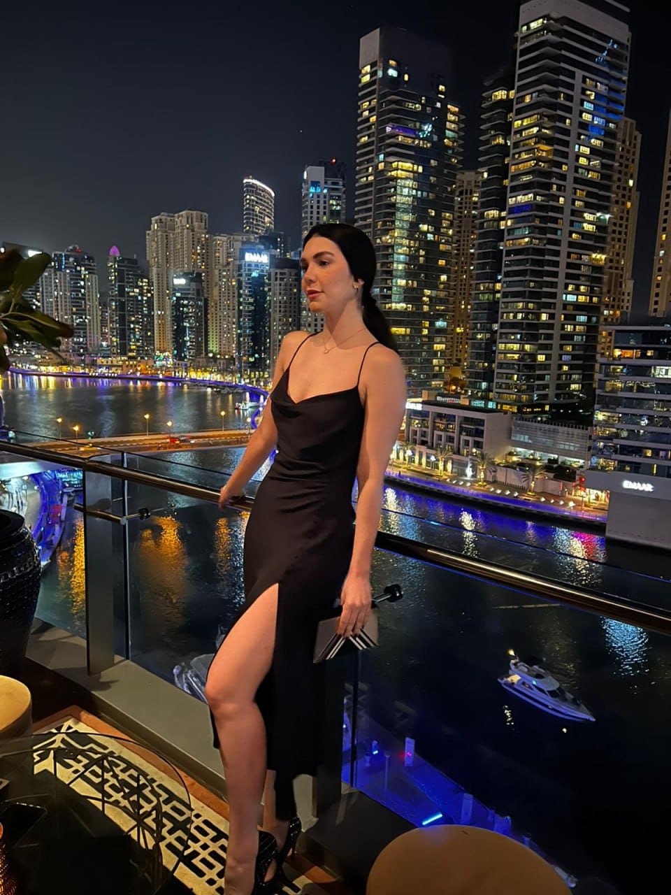 A woman in a black satin slip dress stands on a rooftop terrace at night holding a clutch, city skyline behind.
