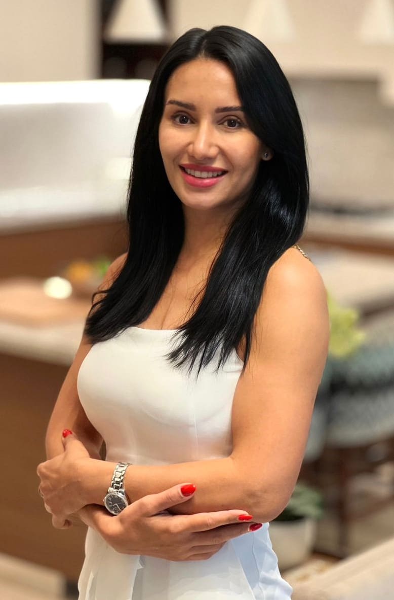 Mona stands with arms crossed, wearing a white sleeveless top and red nail polish, in a softly lit indoor setting.