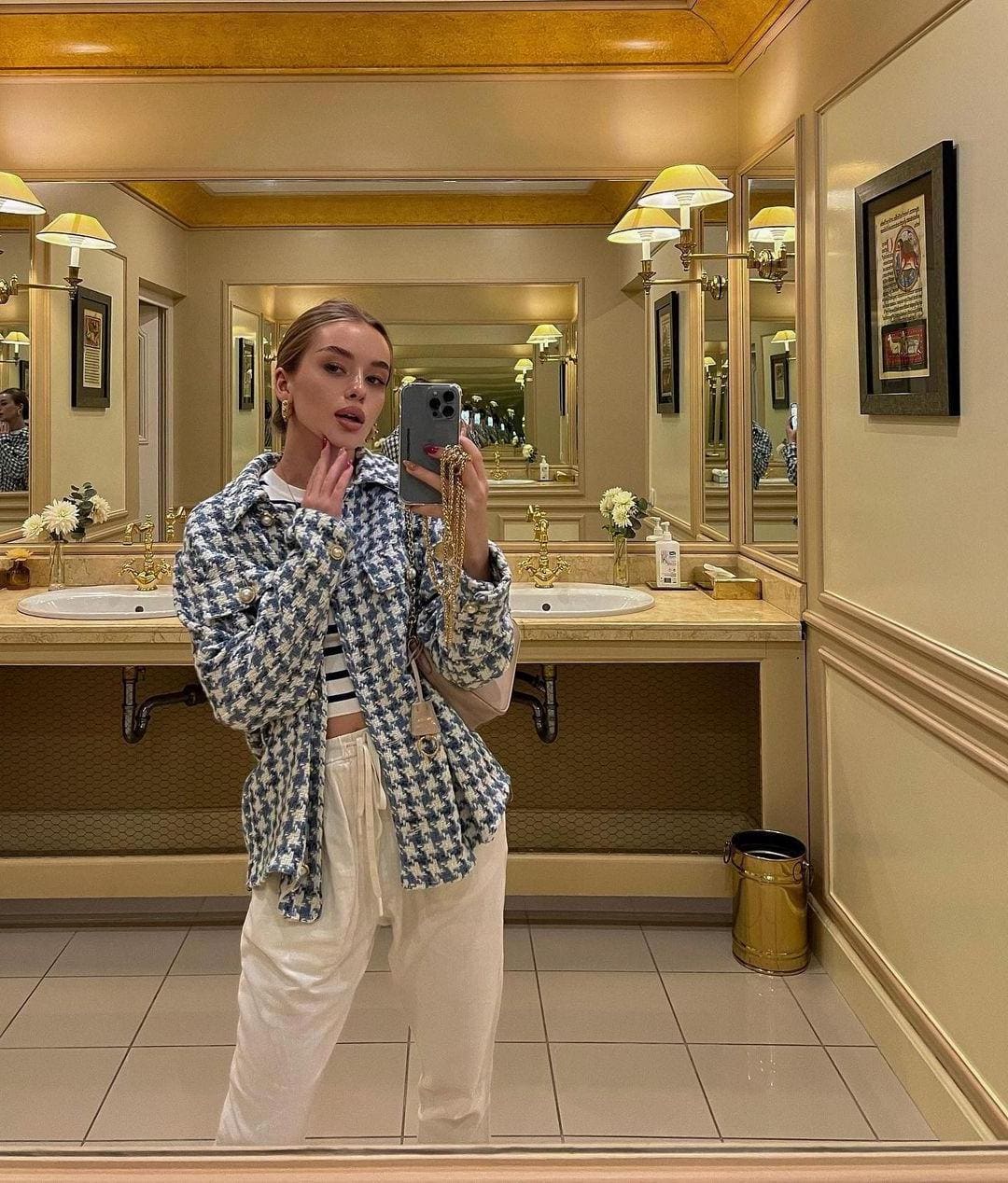 A woman takes a mirror selfie in a gold-toned bathroom wearing a houndstooth jacket, striped crop top, and white trousers.