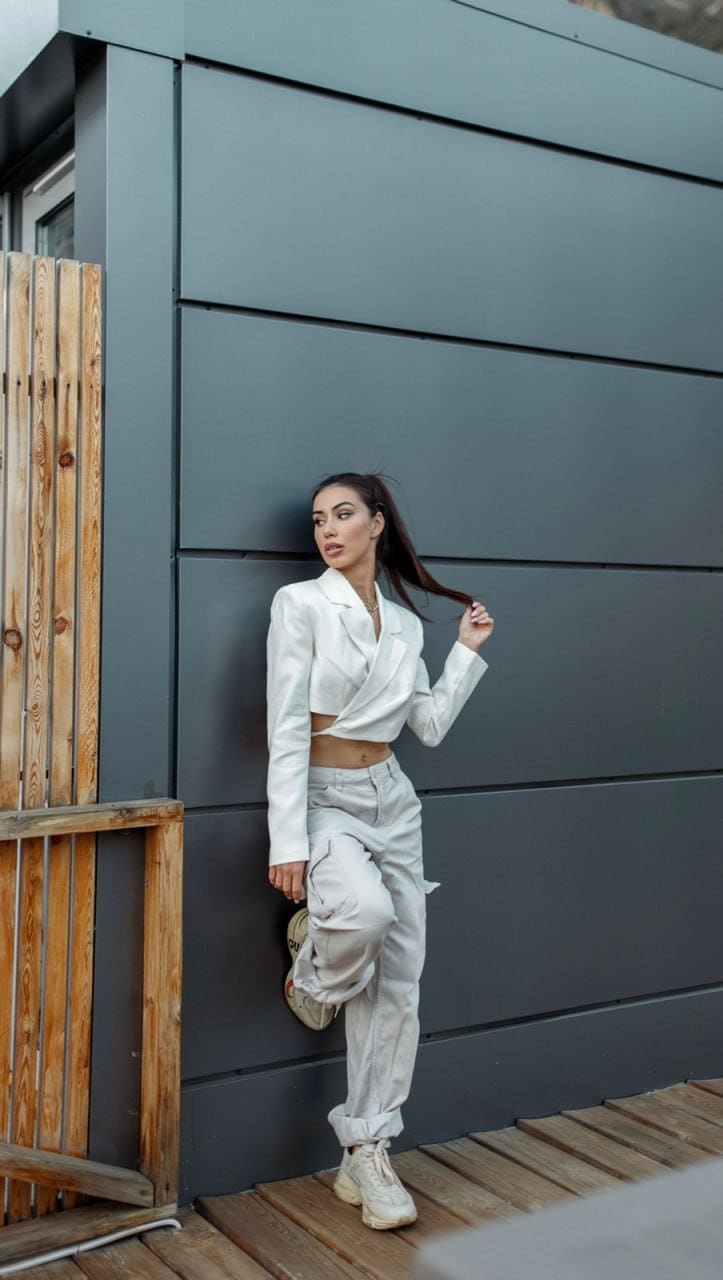 A woman with hair tied back poses on a wooden deck against a gray wall, wearing a white cropped blazer, trousers, and sneakers.