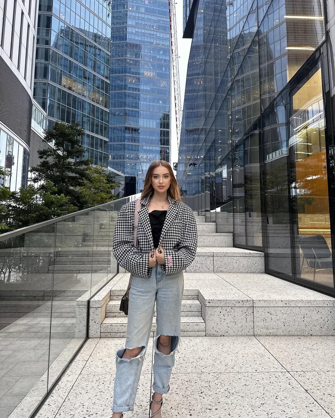 A woman in a houndstooth blazer, ripped jeans, and sandals stands on a stone plaza in front of glass skyscrapers.