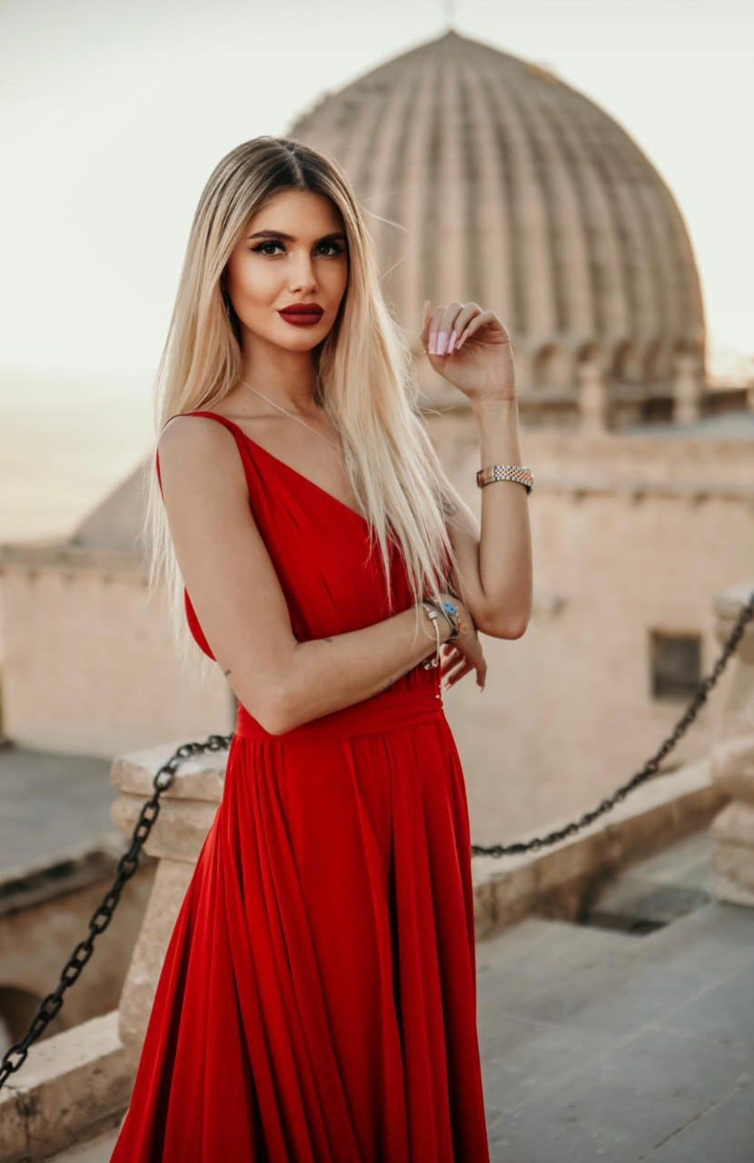 Olivia stands outdoors in a red sleeveless V-neck dress, hand near her face, in front of a domed stone structure.