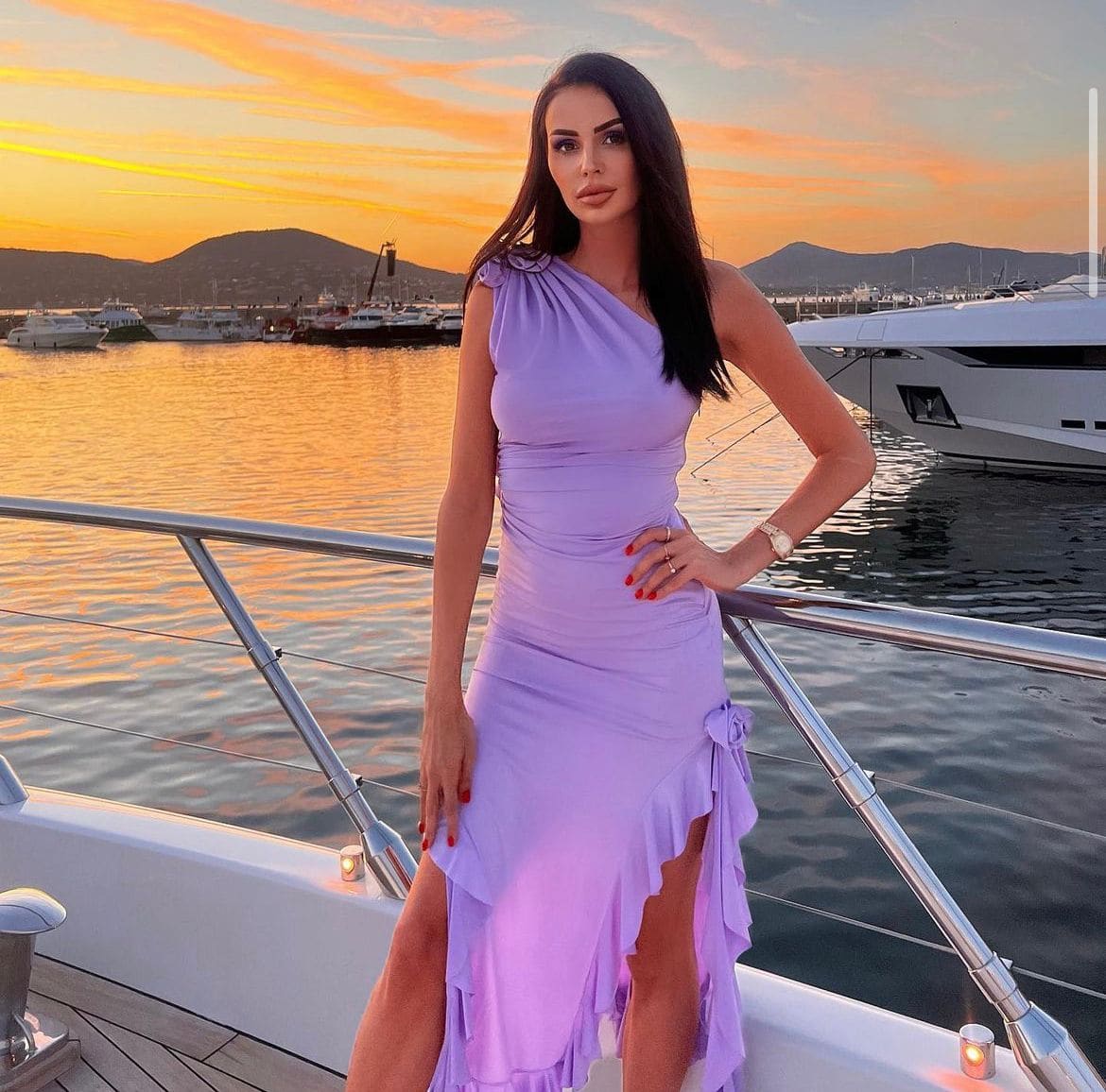 A woman stands on a boat deck at sunset in a lavender one-shoulder dress with a high slit, resting her hand near the railing with yachts behind.