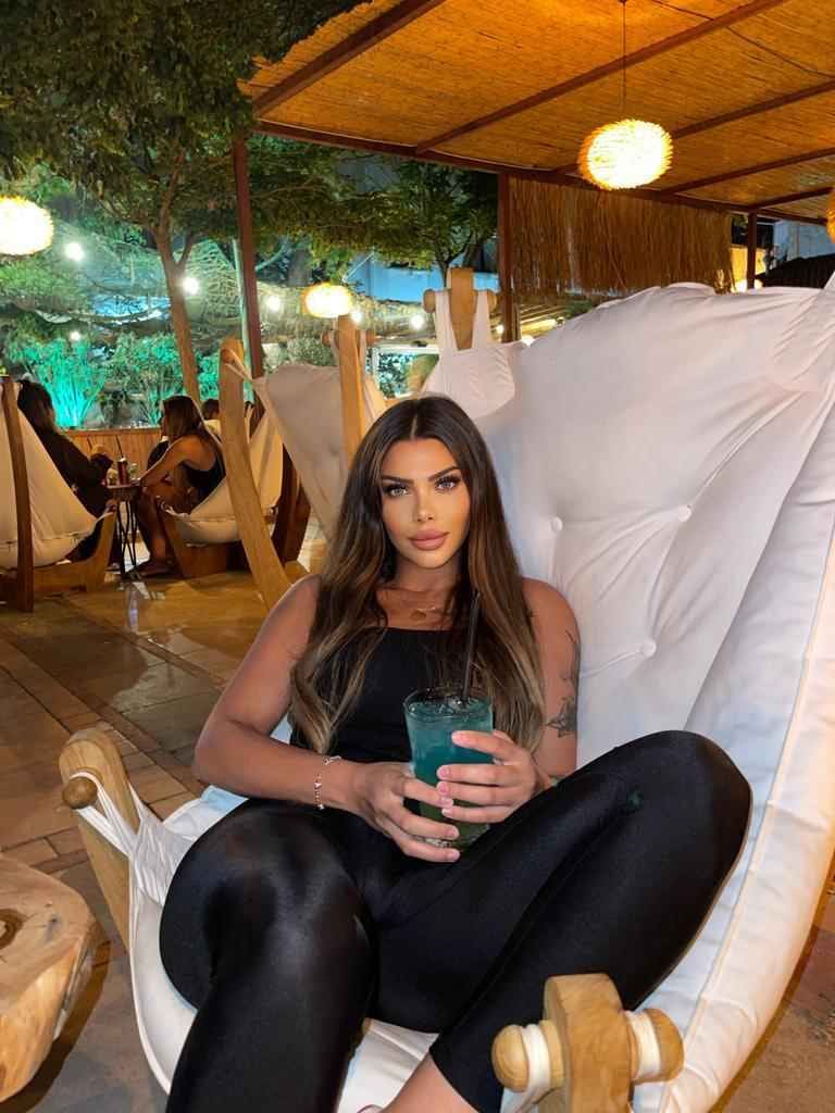 A woman in a black sleeveless top and leggings sits in an outdoor lounge chair, holding a blue drink with a straw.