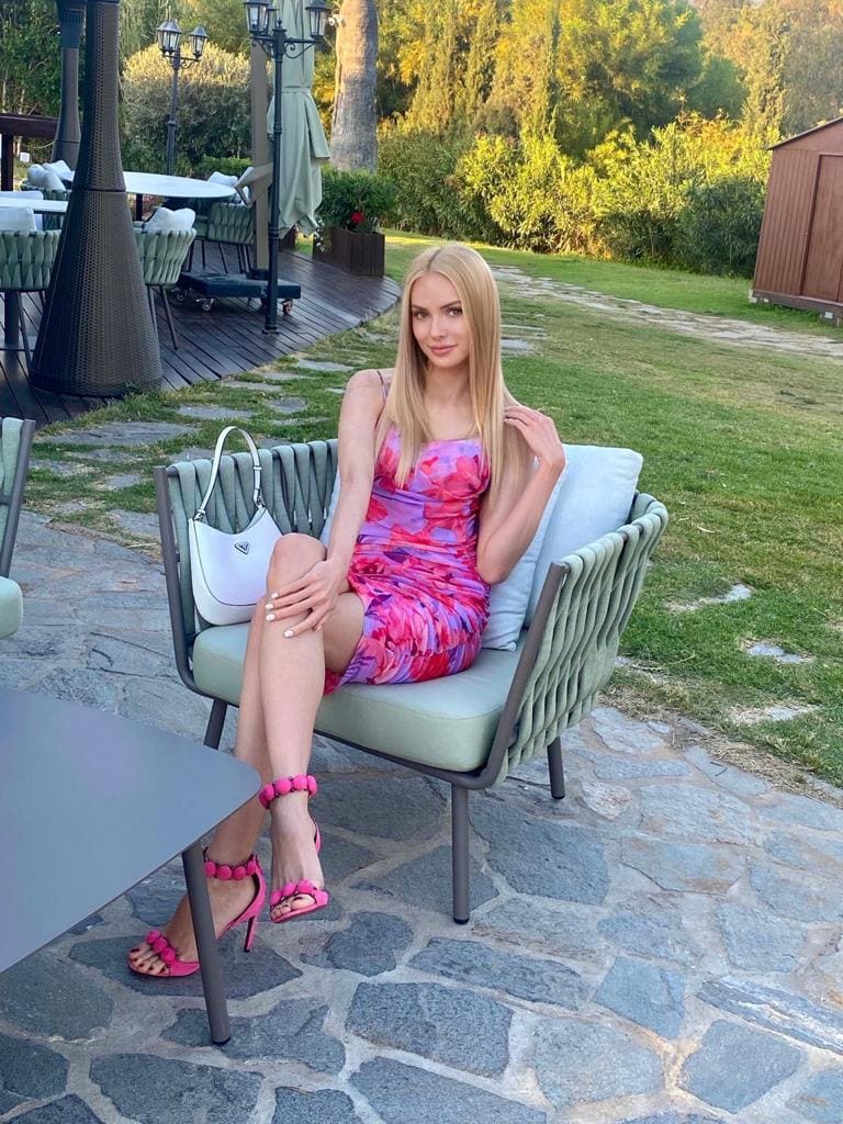 A blonde woman sits in a green outdoor armchair wearing a short pink floral dress and pink heels, with a white handbag nearby.