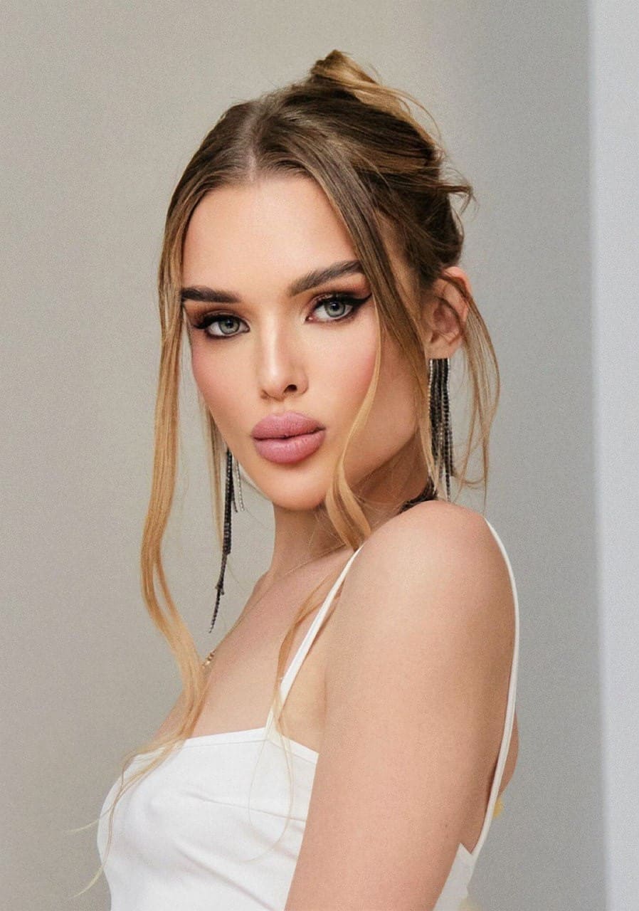 Maria with blonde hair in an updo and earrings wears a white spaghetti-strap top, standing in a three-quarter pose against a light-gray wall.