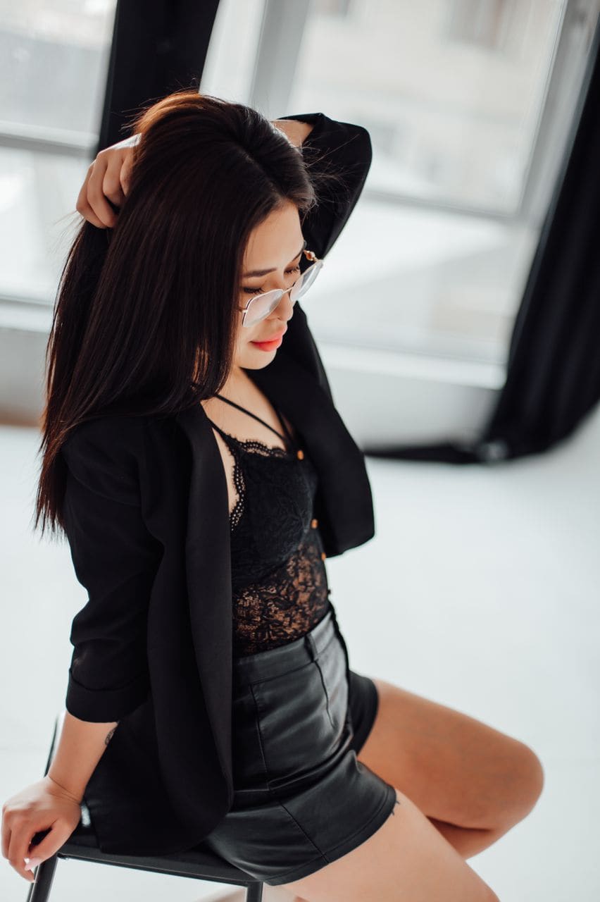 A woman sits in a bright room wearing a black blazer, lace top, and leather skirt, with one hand raised to her hair.