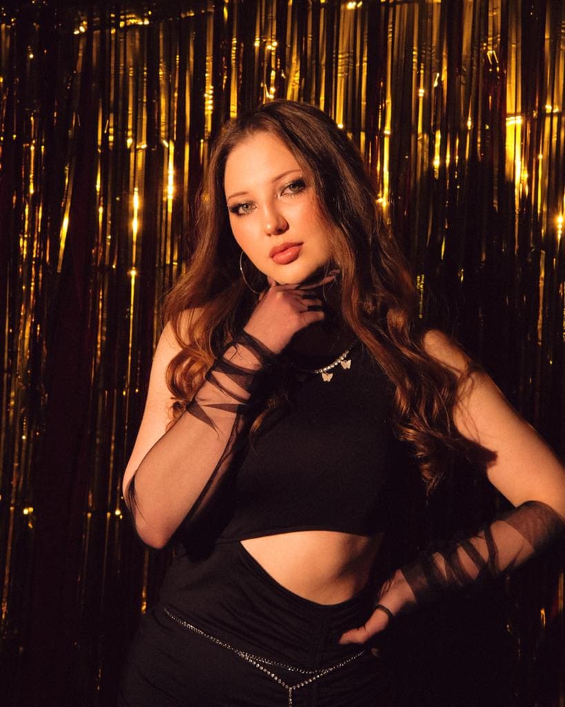 Violetta stands before a gold tinsel curtain in a black cropped top and skirt, hand on neck, other on hip.