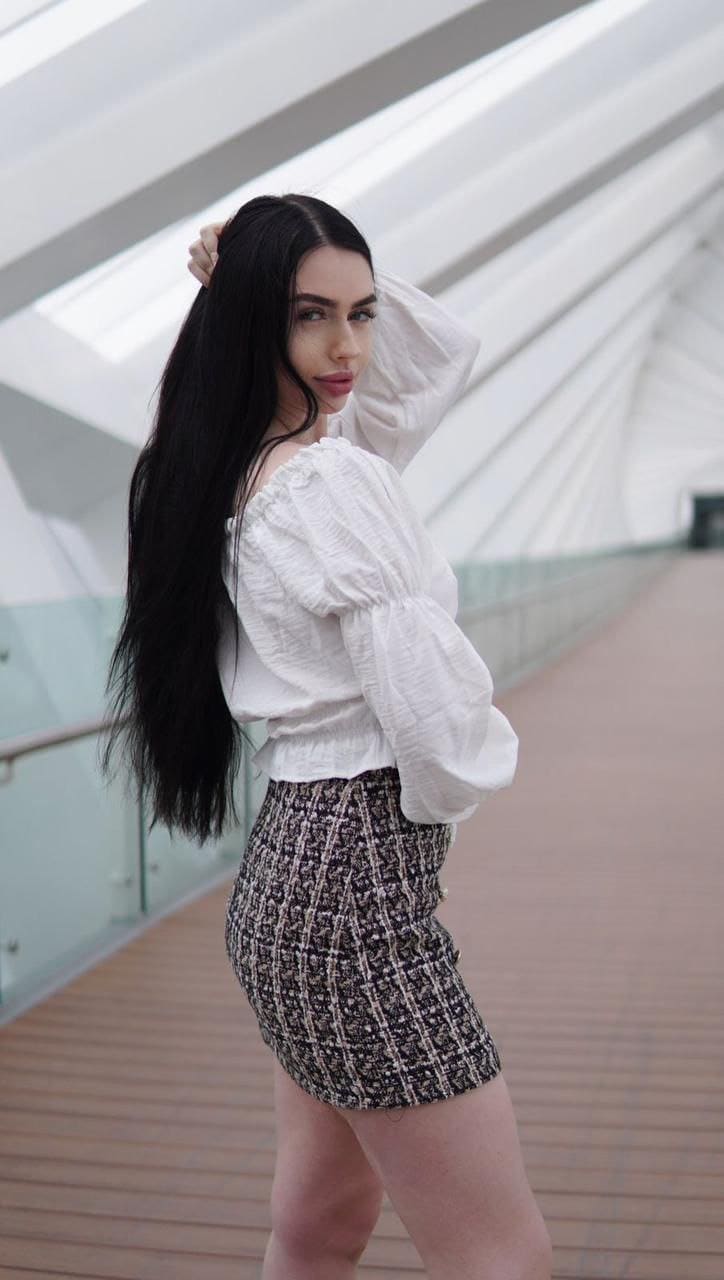 A woman in a white off-shoulder blouse and patterned skirt stands on a wooden walkway, one hand raised to her hair.