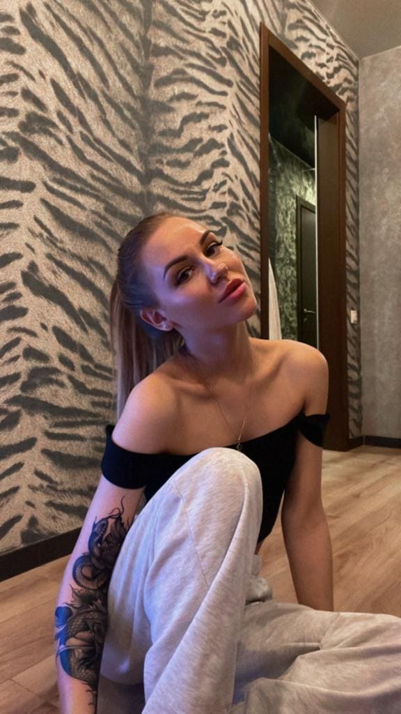 A blonde woman with an arm tattoo sits on a wooden floor in a black off-shoulder top and grey sweatpants against a patterned wall.