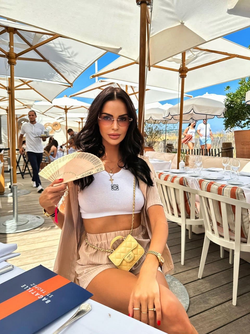 A woman sits at an outdoor table in a white tank top and beige shorts, holding a fan with a yellow crossbody bag at her waist under umbrellas.