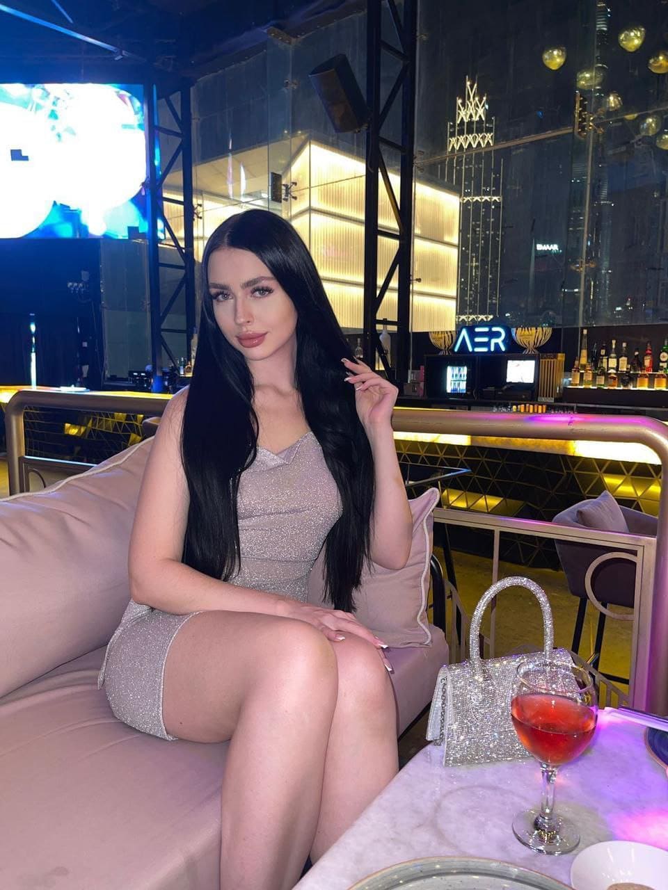 OLGA sits on a pink nightclub sofa in a silver mini dress, hand near her hair, with a red drink on the table beside her.