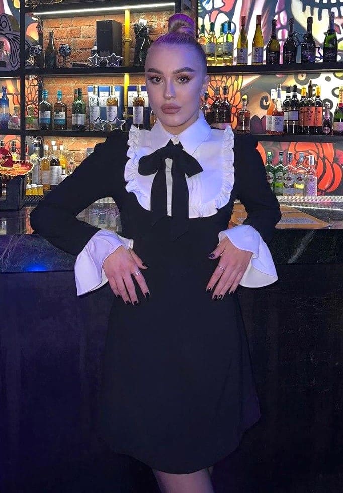 A woman with a purple bun poses in a black dress with a white ruffled collar and bow tie, standing before a bar lined with bottles.