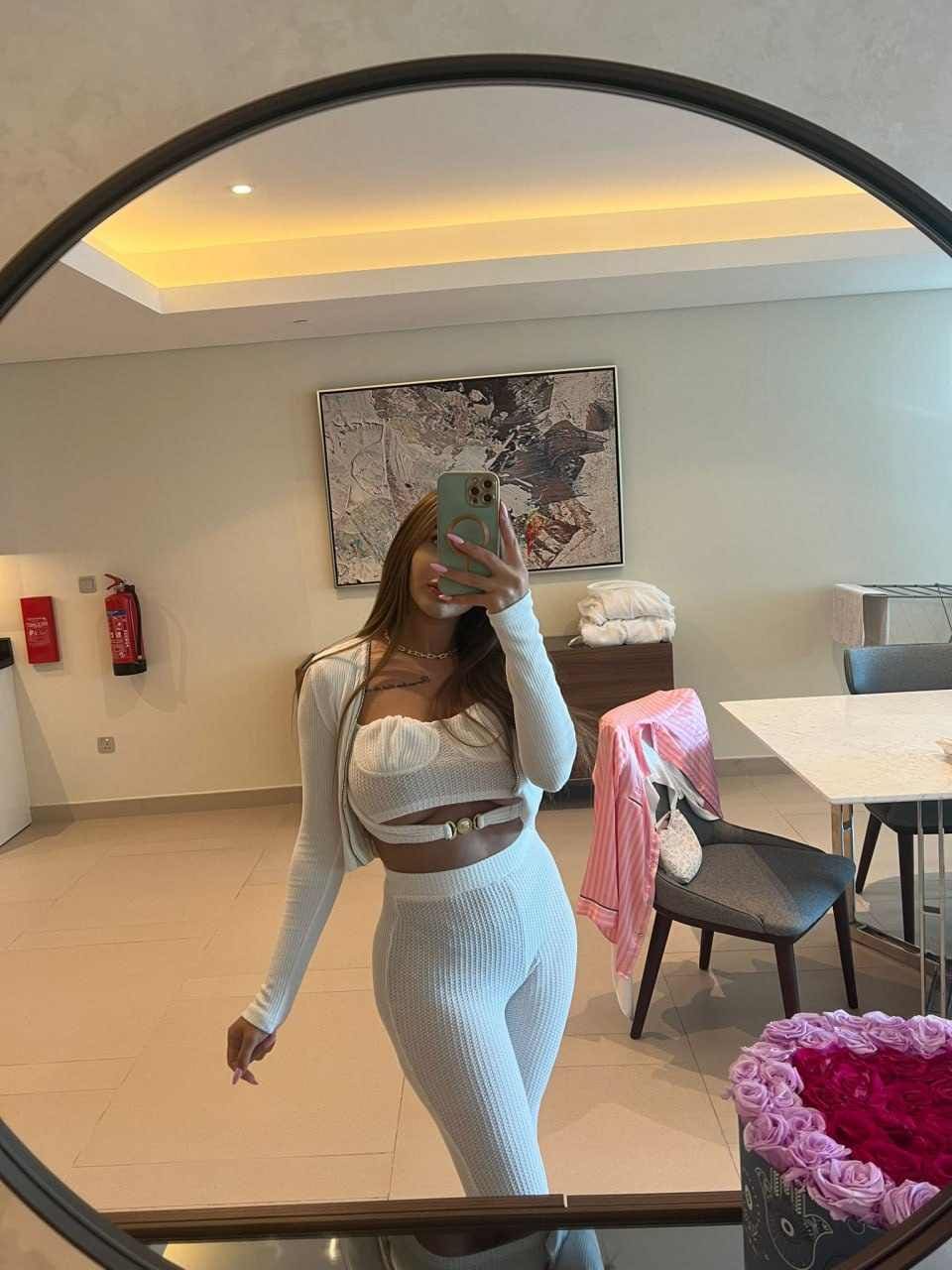 A woman takes a mirror selfie in a white ribbed cardigan, bandeau, and leggings, standing in a room with pink roses in the foreground.