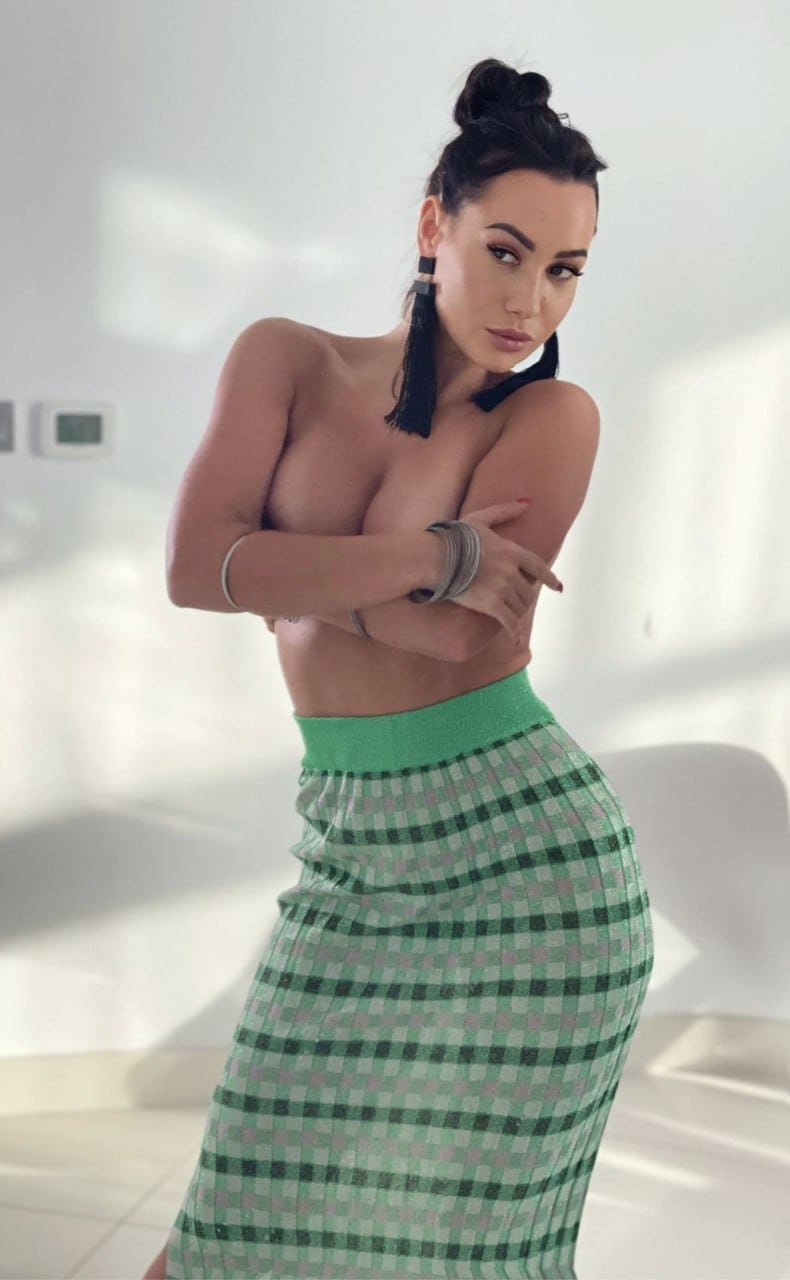 A woman with a high bun stands bare-chested with arms crossed, wearing a green and white checkered midi skirt in a white room.