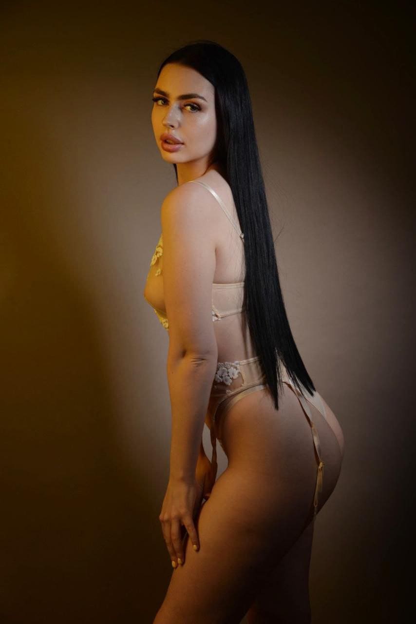 A woman in a beige lace bodysuit with sheer panels and garter straps stands in profile against a plain brown studio backdrop.