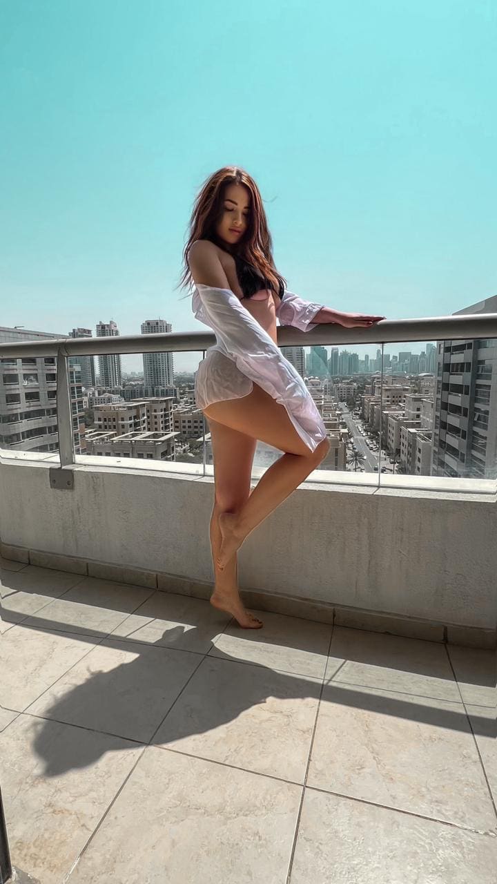 A woman leans on a balcony railing, wearing an unbuttoned white shirt over a pink bikini, with a city skyline behind her.