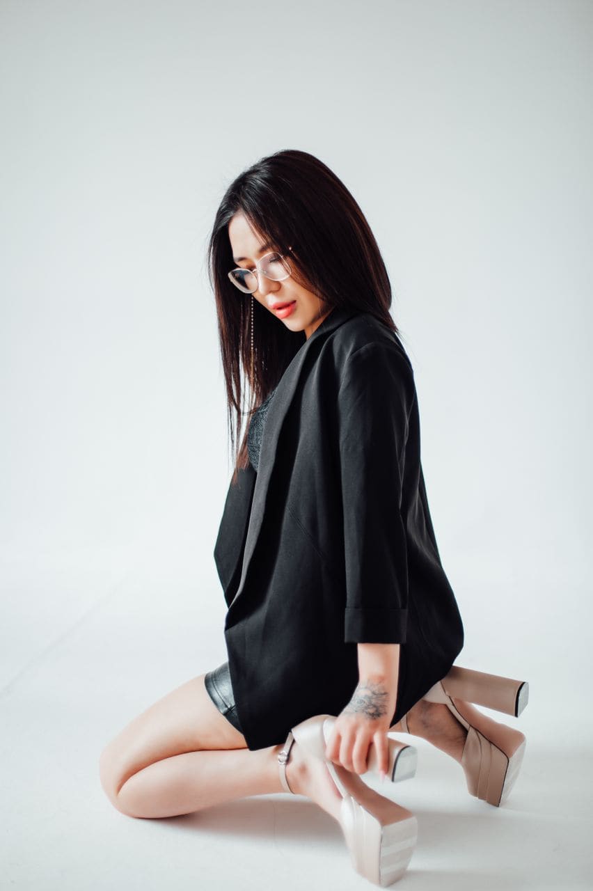 A woman kneels on a white floor in an oversized black shirt and dark skirt, wearing beige platform heels, resting a hand on her foot.