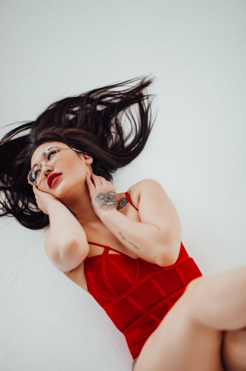 A woman lies on her back on a white surface in a red strappy dress, with one hand near her neck and tattoos visible on her arm.