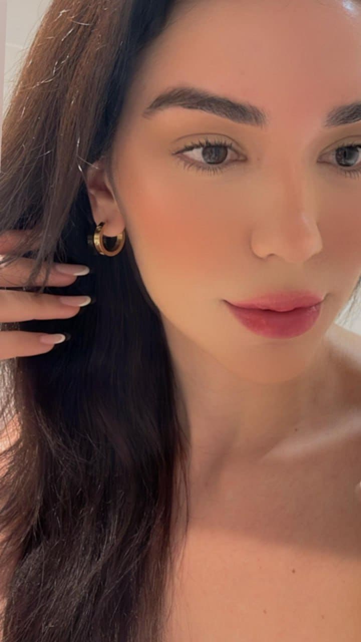 Close-up portrait of a woman with a gold hoop earring, hand near her ear, bare shoulders against a light background.