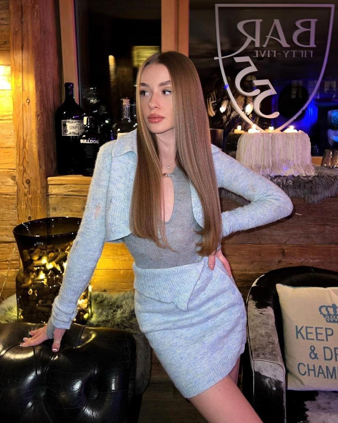 A woman in a light blue knitted cardigan and mini skirt poses in a bar with one hand on her hip and the other on a stool.