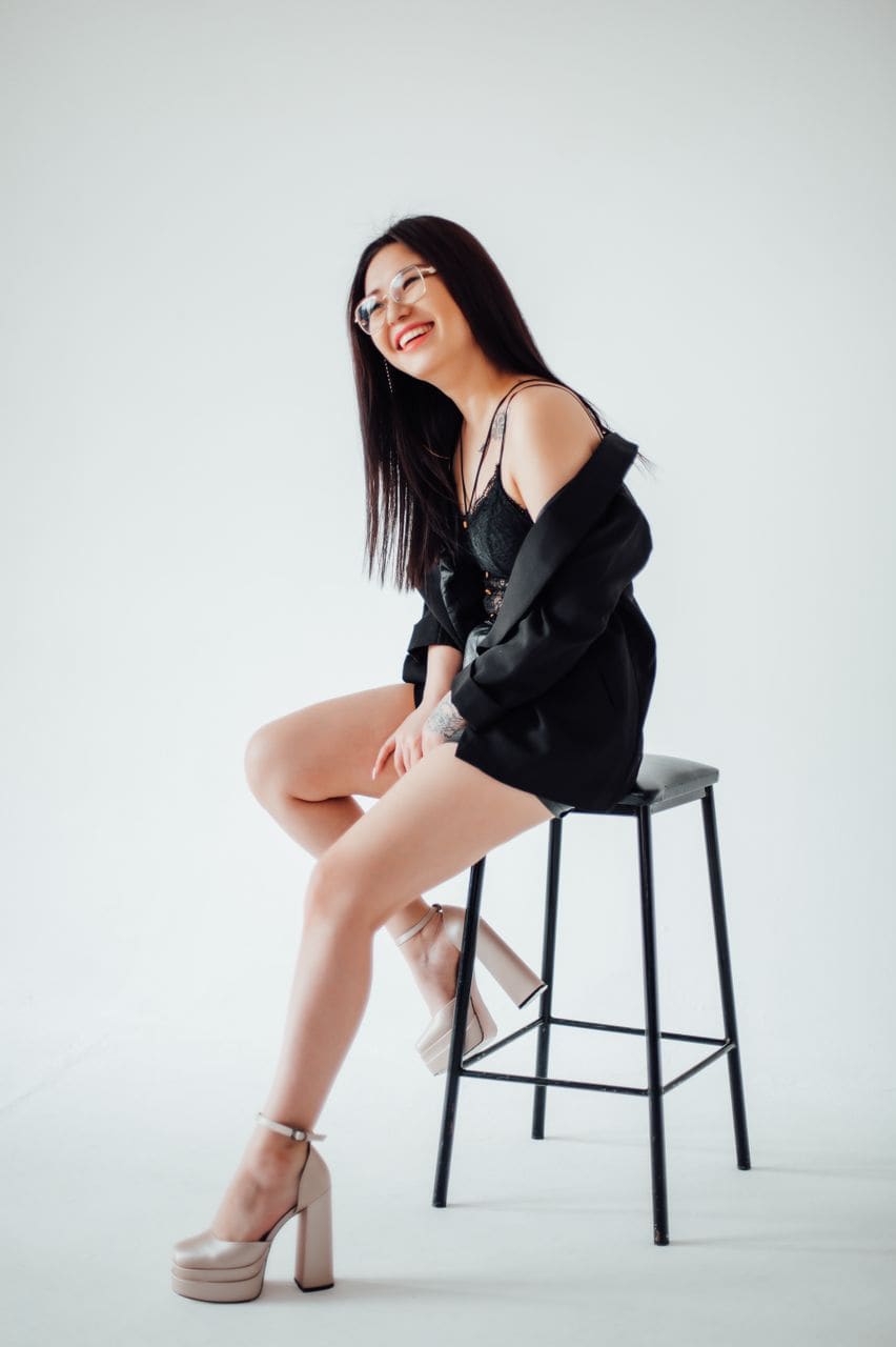A woman sits on a black stool in a white studio, wearing a black lace camisole under an open blazer and nude platform heels.