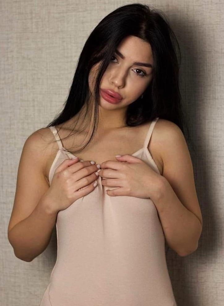 A woman stands against a textured beige wall wearing a light pink spaghetti-strap camisole, holding the fabric at her chest with both hands.