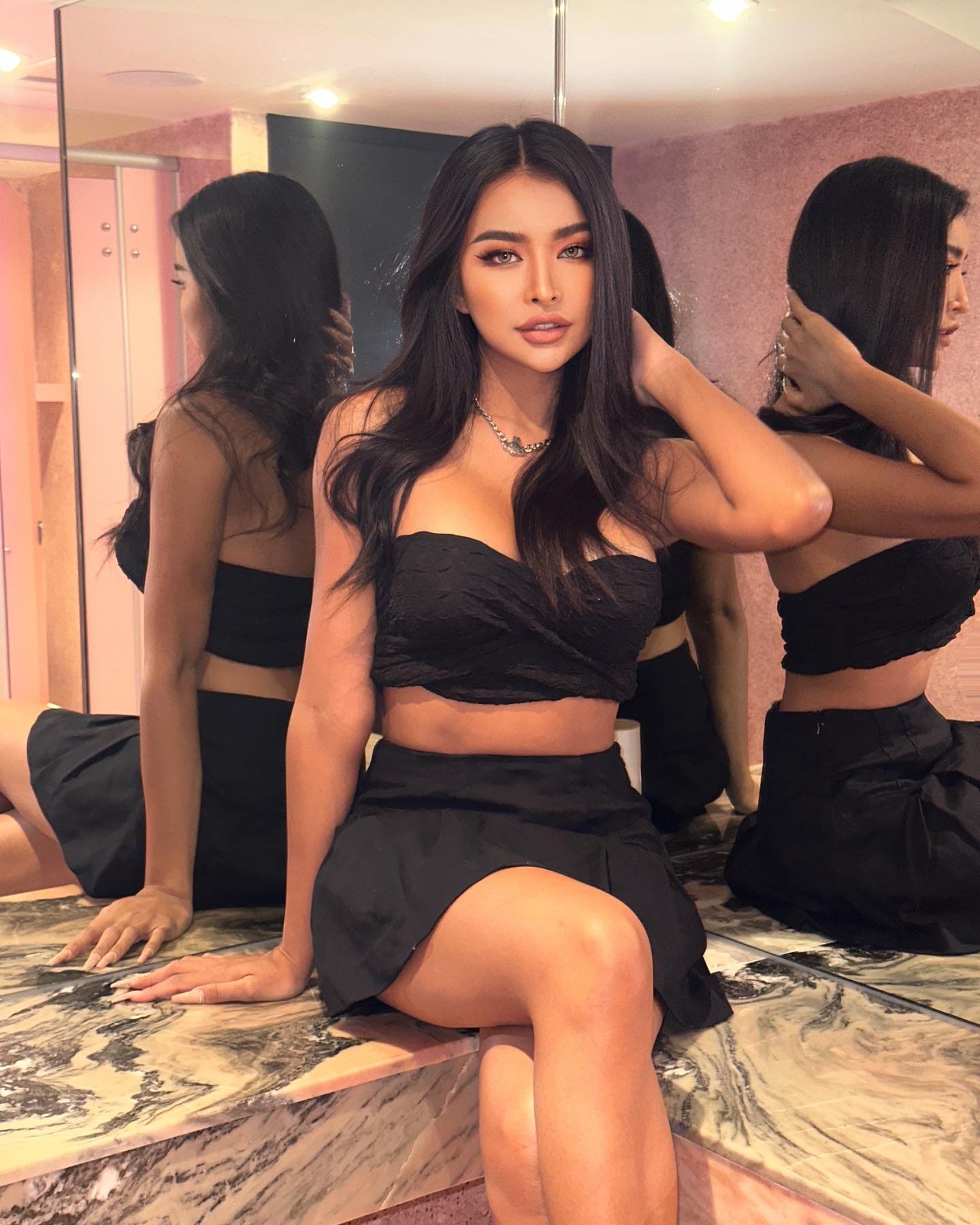 Seated on a marble ledge in a mirrored room, she wears a black strapless crop top and short skirt, one hand on the surface, the other in her hair.