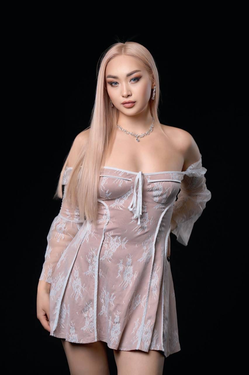 A woman stands in a studio against a black background, wearing a short off-the-shoulder pink lace dress with white trim and a beaded necklace.
