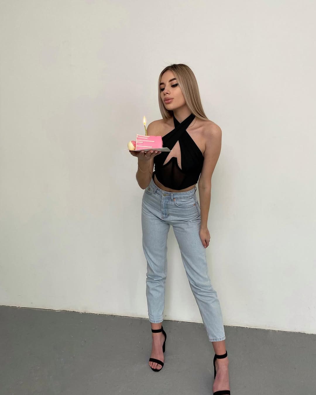 A woman in a black halter top and light-wash jeans stands against a white wall, holding a pink cake with a lit candle on a plate.