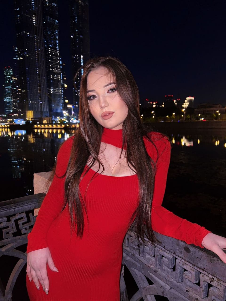 A woman stands by a waterfront railing at night in a red long-sleeved dress, with illuminated buildings and a bridge behind.