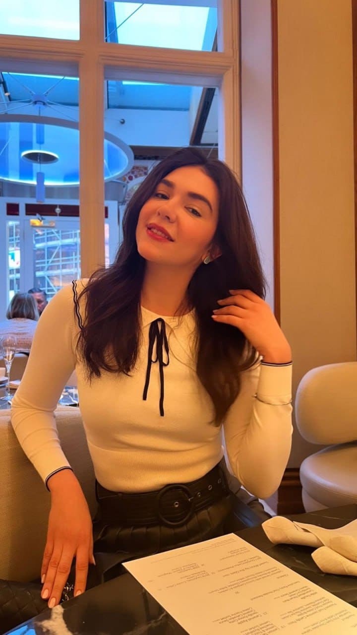 Beatriz sits at a restaurant table in a white top with a black tie and belt, one hand touching her hair.