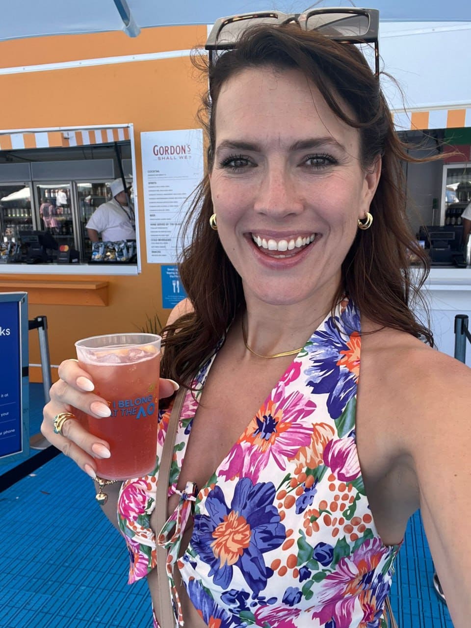 Nicole holds a pink drink in front of an orange and white food stand, wearing a floral halter top and sunglasses on her head.