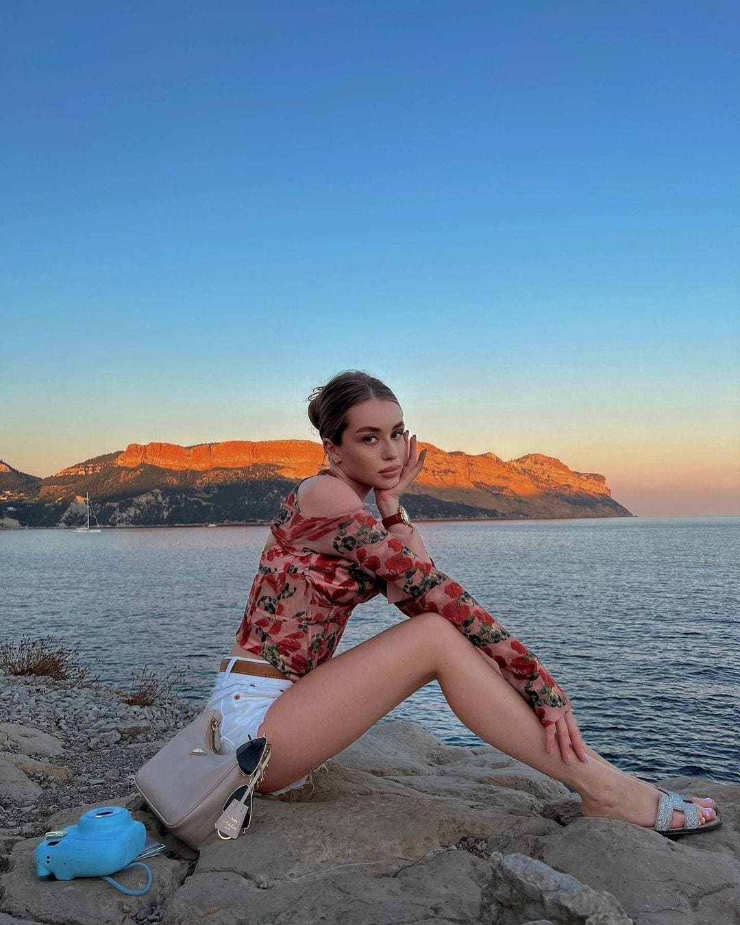 A woman sits on a rocky shore at sunset wearing a sheer floral blouse, white shorts, and strappy sandals with a camera nearby.