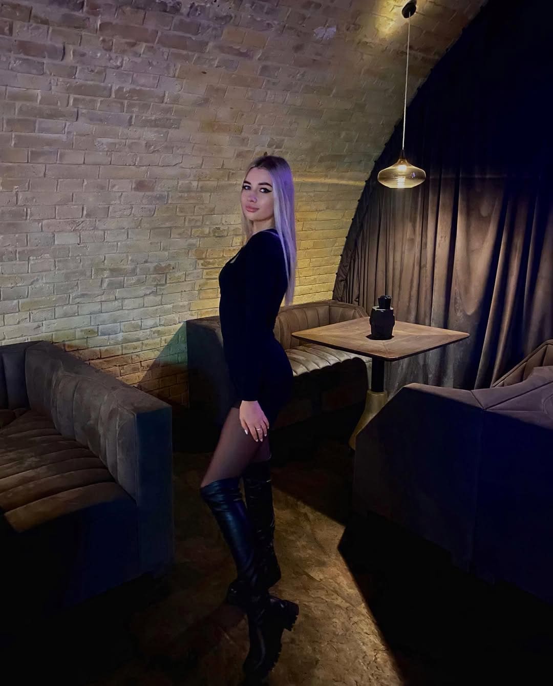A woman with light-purple hair stands in profile in a dim room with brick walls, wearing a black mini dress and knee-high boots.