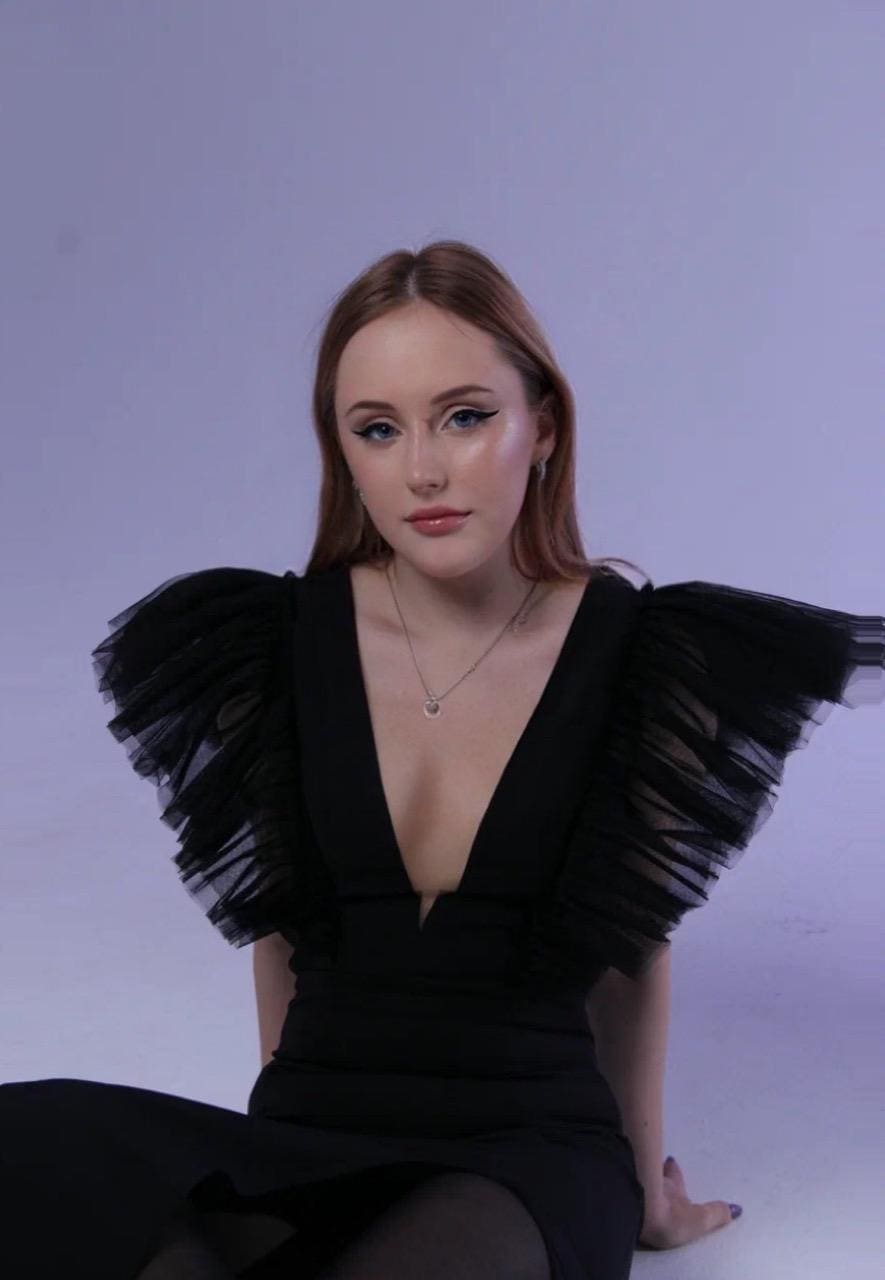 Annabella sits on a white floor against a light background, wearing a black V-neck dress with ruffled tulle sleeves and a thin necklace.