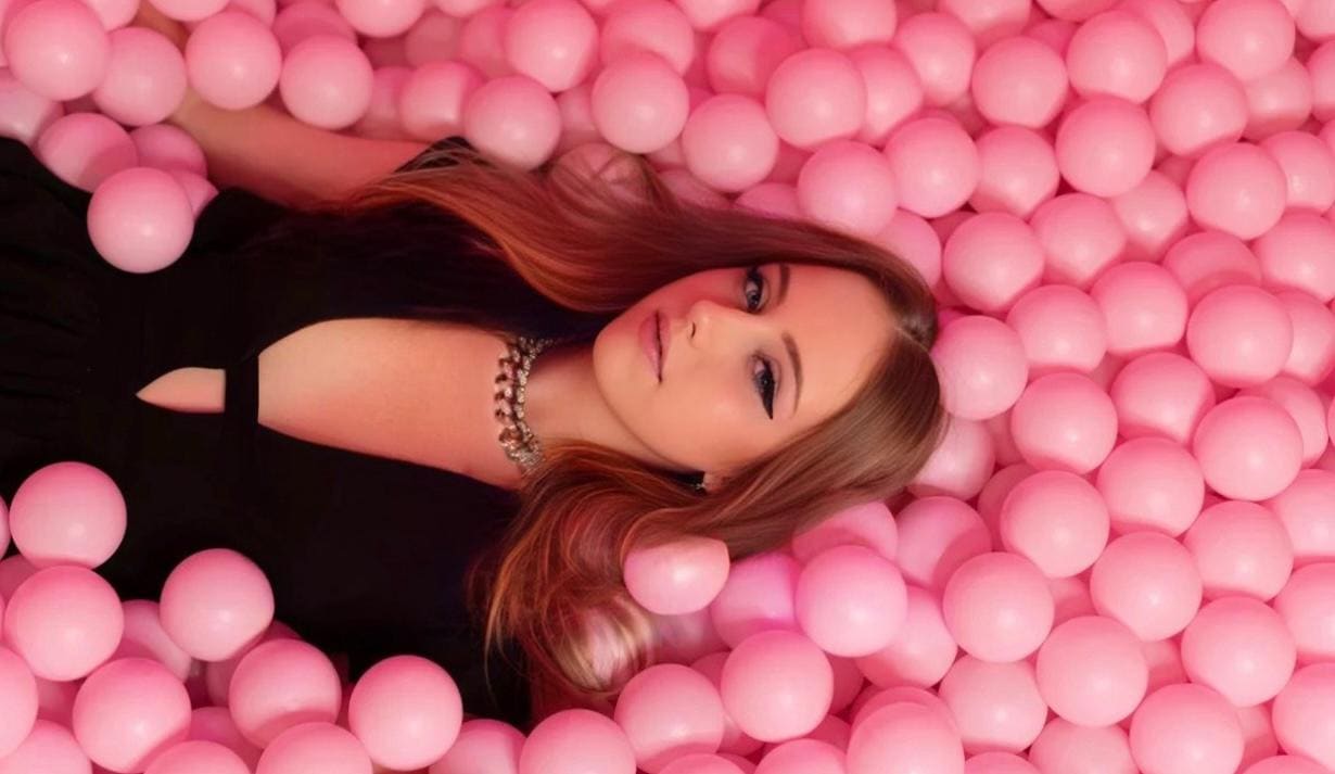 Lying on her side in a pit of pink plastic balls, she wears a black sleeveless keyhole-neck top and beaded necklace, looking at the camera.