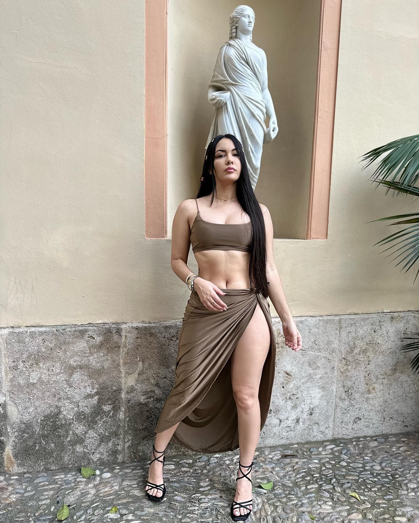 A woman in a brown crop top and high-slit wrap skirt stands on pebbles near a beige wall with a statue in a niche.
