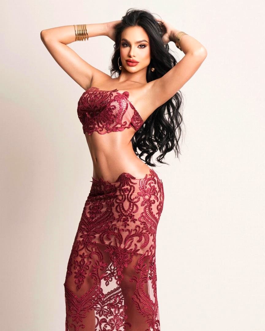 Standing with hands behind her head in a burgundy lace bra top and sheer high-waisted skirt against a plain light background.