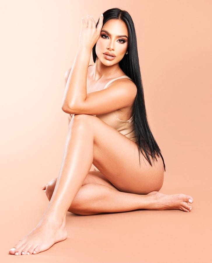 Sitting on the floor in a beige one-piece swimsuit with one hand raised to her head against a plain peach background.