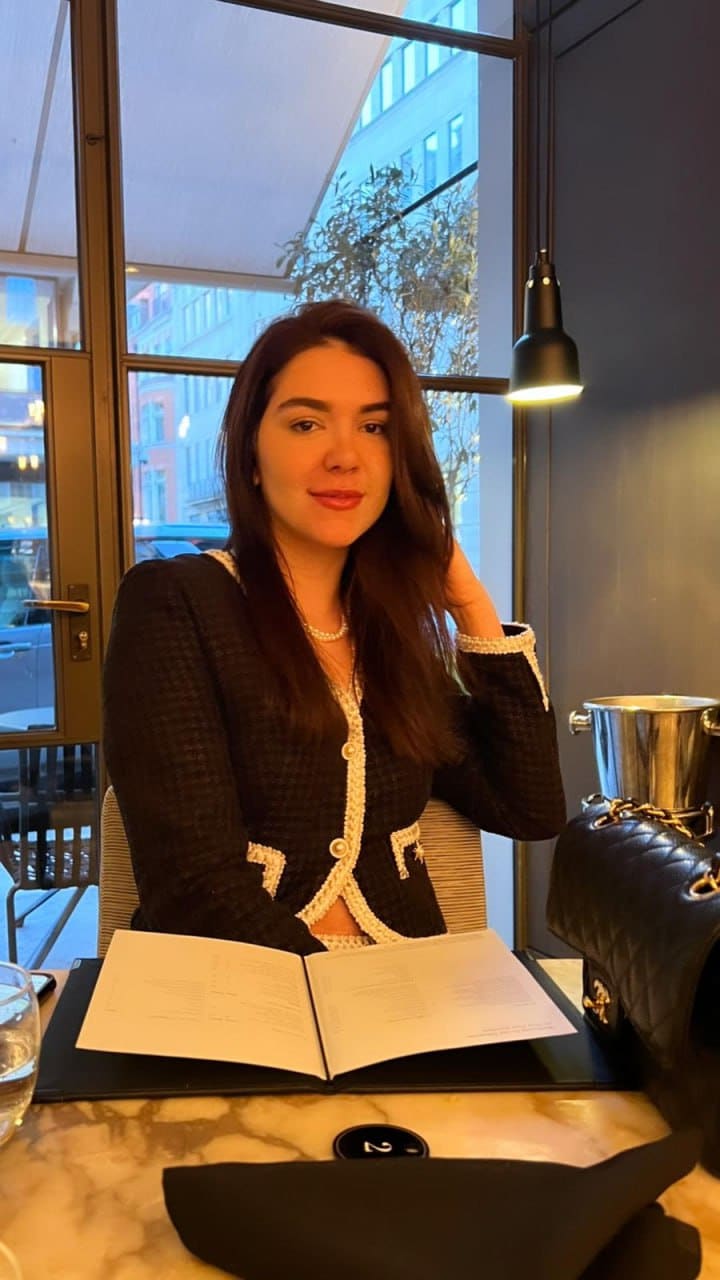 A woman in a tweed jacket and pearl necklace sits at a marble restaurant table with an open menu and window view.