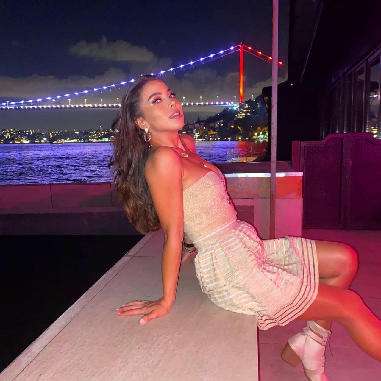 A woman sits on a stone ledge at night in a textured strapless top and striped mini skirt, with a lit suspension bridge and water behind.