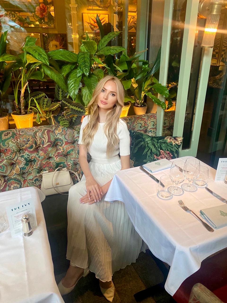 A blonde woman sits at a restaurant table in a white top and pleated skirt, with a white handbag on the sofa beside her.