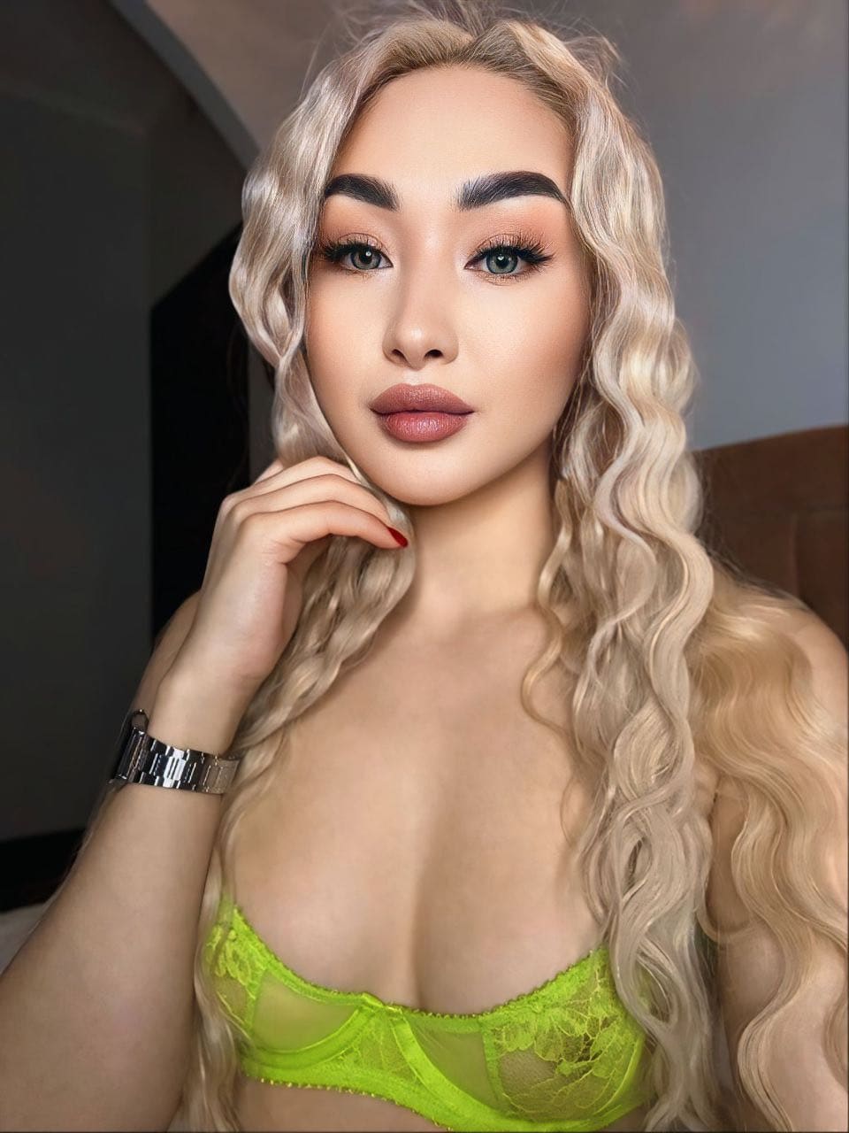 A woman in a lime-green lace bralette and metal wristwatch poses with one hand near her chin in a dimly lit indoor setting.