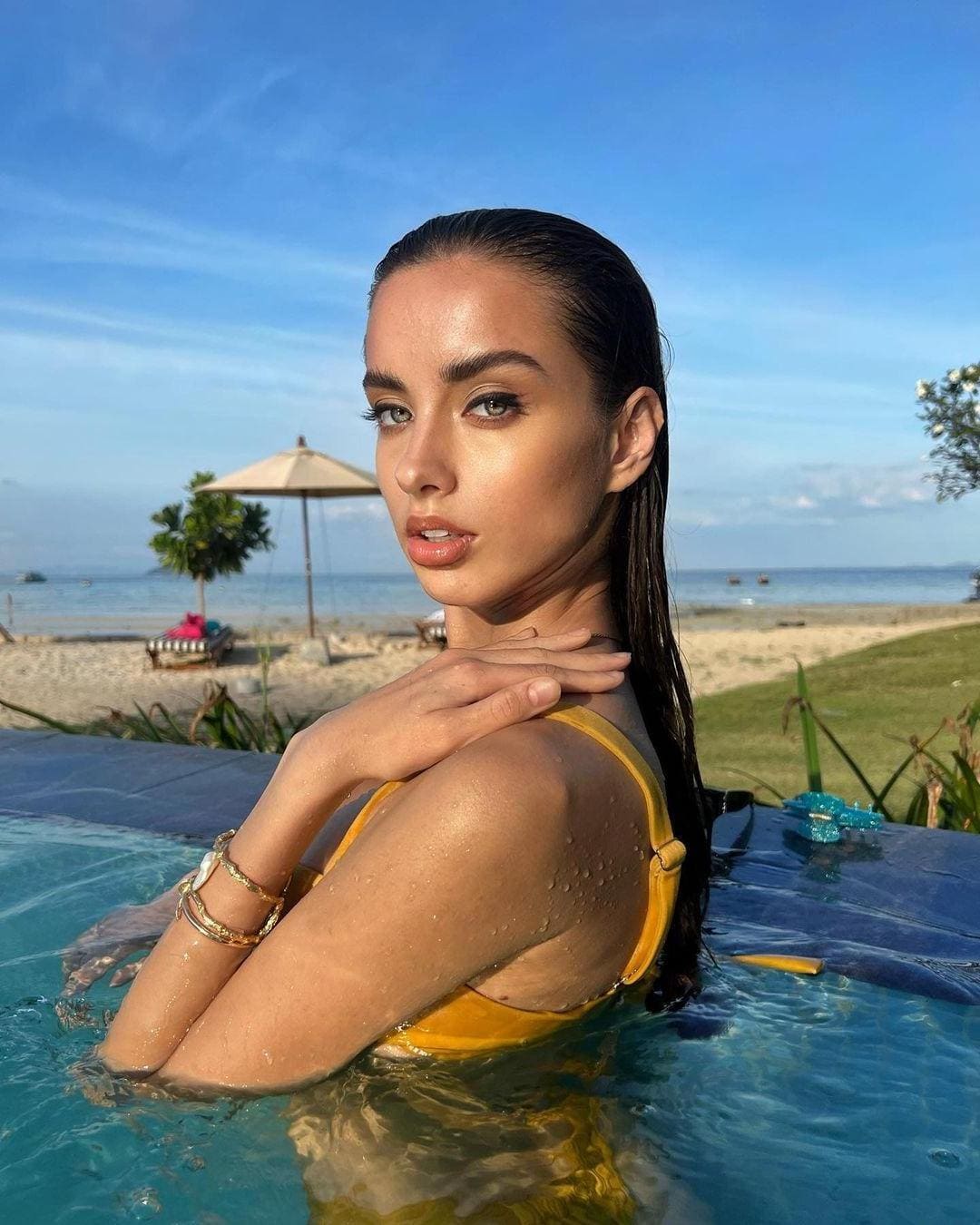 A woman with wet hair sits in a pool wearing a yellow one-piece swimsuit, her hand on her shoulder, against a beach and blue sky backdrop.