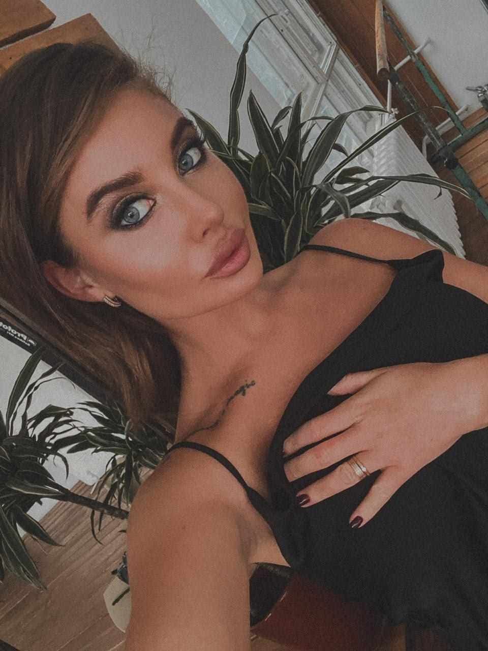 Aleksa wears a black spaghetti-strap top with a deep neckline, hand on chest, posing indoors among potted plants and wooden furniture.