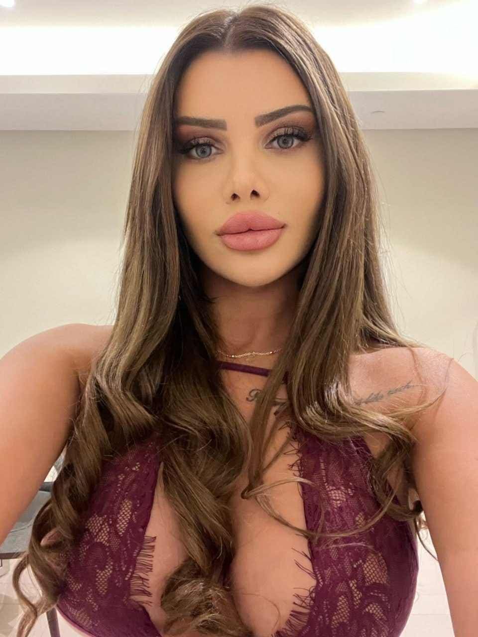 A woman poses facing the camera in a deep-plunging burgundy lace top and choker, set against a plain light-colored interior wall.
