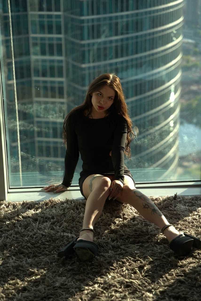 Klava sits on a brown rug by a window, wearing a black top and skirt, with tattoos on her legs and a skyscraper visible outside.