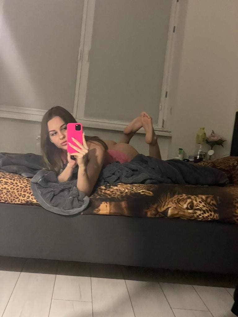A woman in a pink top lies on a leopard-print bed with a grey blanket, holding a pink phone while raising her bare feet.